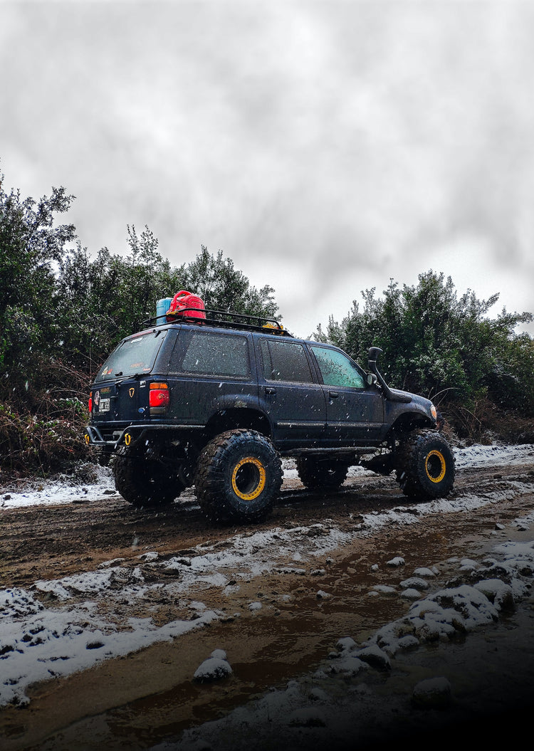 Jeep in falling snow on dirt road with scrub brush behind.