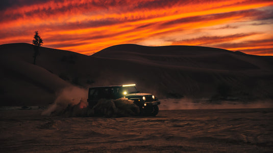 Jeep moving quickly through sand. Vibrant sunset behind. Photo by Abed Ismail.