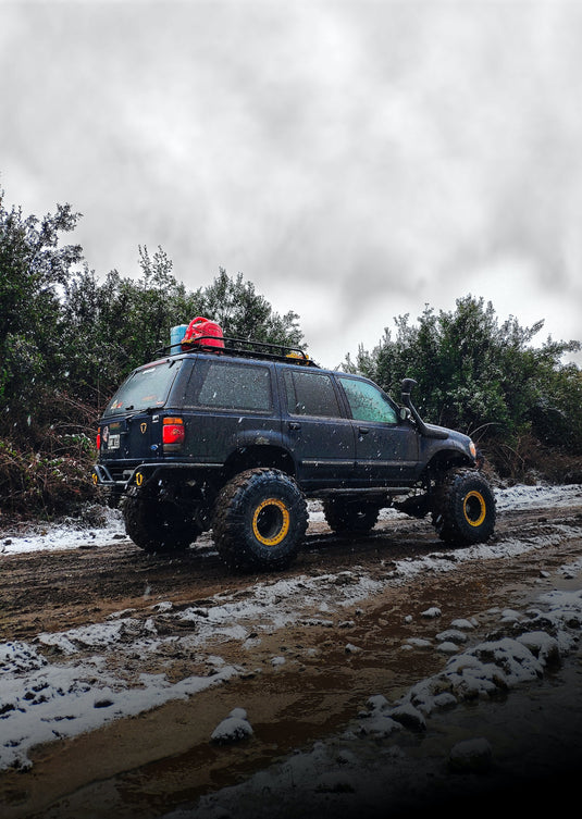 Jeep in falling snow on dirt road with scrub brush behind.